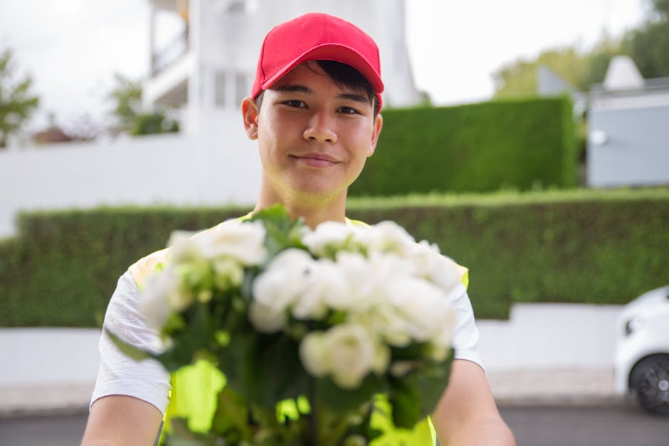 A delivery person wearing a red cap and reflective safety vest is smiling as they hold a bouquet of fresh white flowers with green foliage. The bouquet appears to be arranged with a variety of flowers