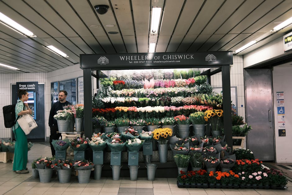 A flower stall in a subway station operated by Wheeler of Chiswick, featuring a variety of fresh flowers arranged in bouquets and buckets. The arrangement includes vibrant blooms such as white, pink, 