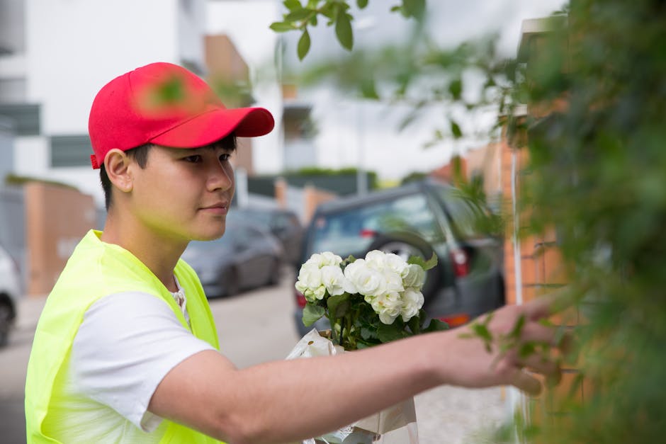 A young male flower delivery courier wearing a red cap and a neon yellow vest is seen placing a bouquet of white hydrangeas and roses against a green leafy hedge in an outdoor residential area. The bo