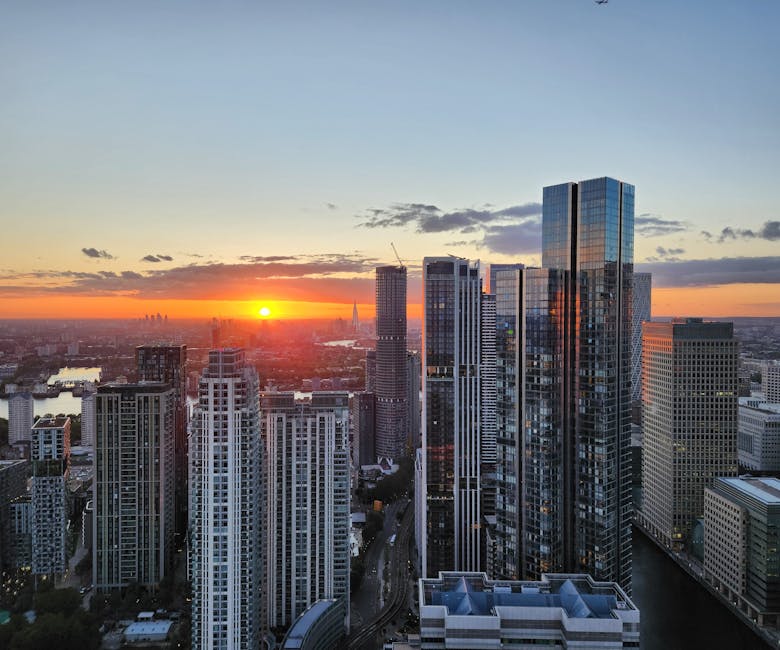 A panoramic view of a London cityscape at sunset, featuring tall modern skyscrapers with glass facades reflecting the warm orange and yellow hues of the setting sun. In the foreground, several high-ri