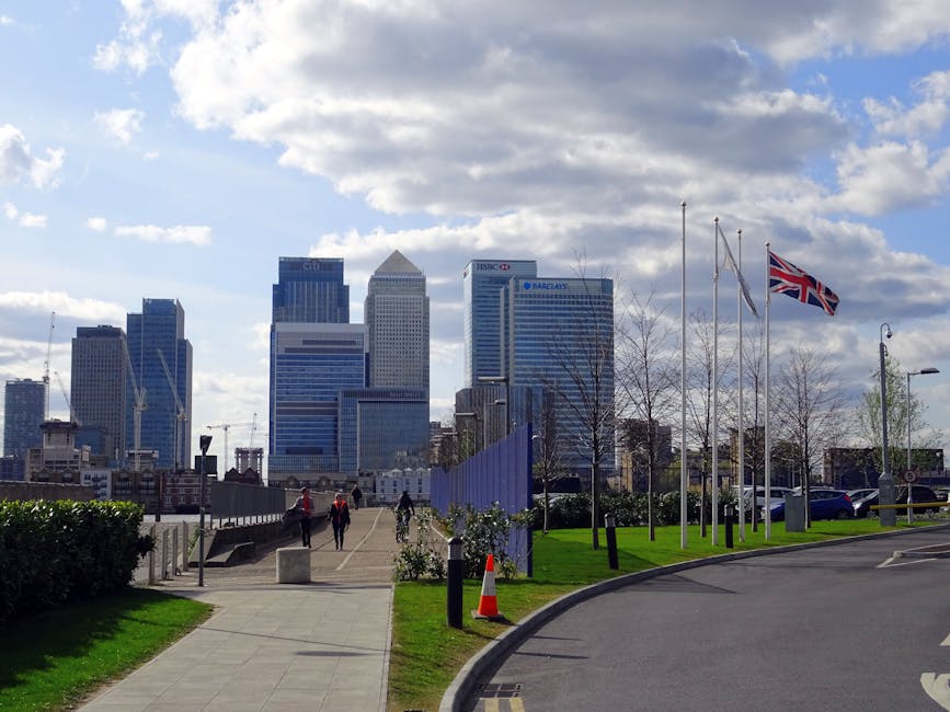 A view of the Canary Wharf skyline in London E14 during daytime, featuring modern glass skyscrapers with blue and silver facades under a partly cloudy sky. In the foreground, a paved promenade runs pa