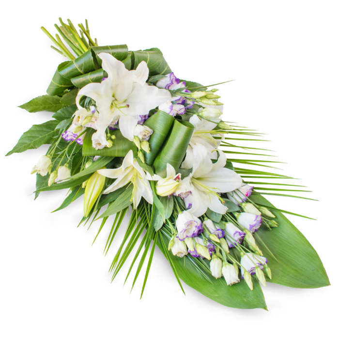 An elegant funeral and memorial spray is laid out diagonally against a crisp white backdrop, its long, tapered outline suggesting a gentle sigh or a single falling petal. The composition begins with a lush gathering of white oriental lilies at the upper left, where the stems converge. Some lilies are fully open, revealing smooth, outward-curving petals and subtle stamens at their centre, while others are partially open or held in sleek buds, offering varied heights and forms that guide the eye along the arrangement. Interwoven between the lilies are lisianthus flowers, predominantly white yet softly rimmed with lavender or pale purple, adding a delicate, painterly contrast. Numerous buds of lisianthus, still tightly furled, introduce a sense of depth and quiet potential, as though the spray will continue to unfold in the days following the service. Surrounding these blooms is a carefully crafted foundation of green foliage: broad, dark leaves provide a rich, solid base, and several have been meticulously folded or rolled into sculptural accents, lending a contemporary touch to the otherwise classic form. From the right-hand side and trailing to the bottom tip, long, slender palm fronds extend outward in gentle arcs, reinforcing the elongated teardrop shape and giving the piece a sense of poised movement. The white and green palette, punctuated by just a whisper of lavender, evokes stillness, purity, and respectful remembrance, perfectly suited for funeral services, wakes, or memorials in Poplar and nearby riverside communities. Thoughtfully designed by local florists at Flower Delivery Poplar, the spray feels like a soft-spoken gesture of sympathy, bringing an atmosphere of calm grace to any farewell setting.